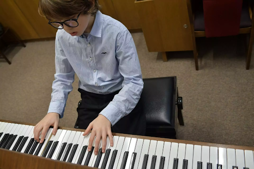 Young boy wearing glasses playing the piano attentively during a music lesson in a classroom setting.